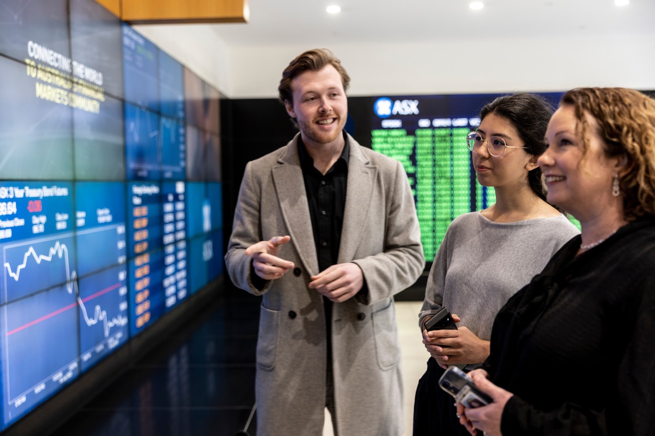 Three people viewing electronic market data displays and charts on large digital screens.