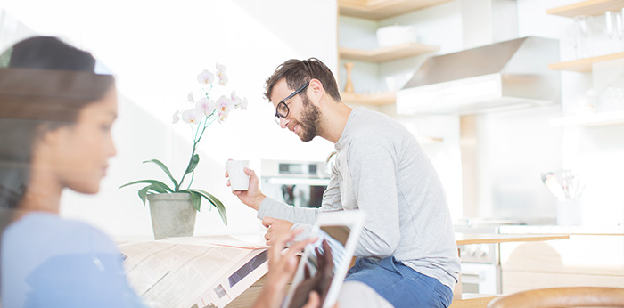 Two people using digital devices in a bright kitchen area, with one person holding a tablet and another seated nearby holding a mug.