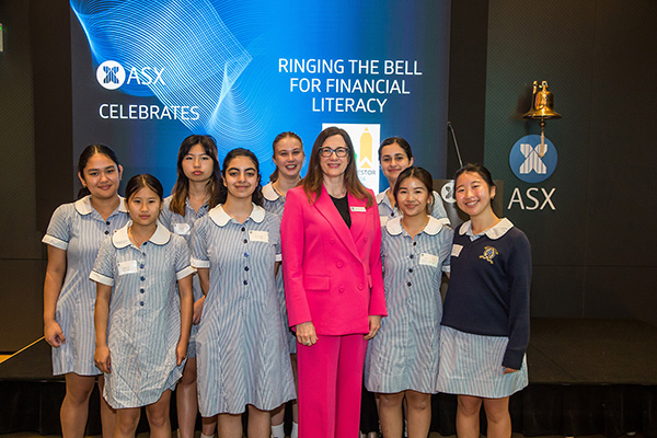 Group photo at an ASX event focused on financial literacy. ASX CEO Helen Lofthouse stands in a bright pink suit stands with students in school uniforms. The backdrop features the ASX logo and the phrase 'Ringing the Bell for Financial Literacy.' A ceremonial bell is visible on the right.