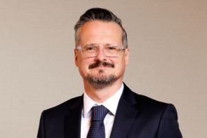 Head-and-shoulders professional portrait of Jamie Crank wearing a suit jacket, facing the camera against a plain light background