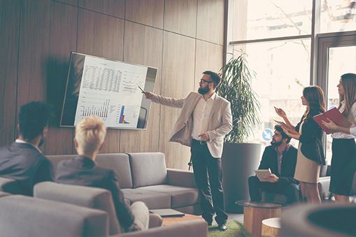 Group of people seated indoors while one person gestures toward a presentation screen