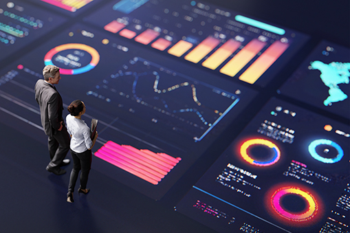 Two business professionals stand on a dark, expansive surface, examining large digital dashboards projected around them. 