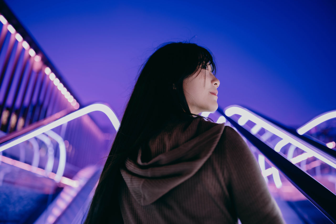 A young woman with long dark hair stands on an outdoor staircase at night, looking off into the distance. 