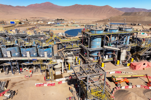 Industrial mining and mineral processing facility with pipes, tanks, and machinery in a desert landscape surrounded by mountains.