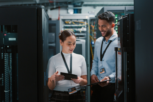 Two data centre technicians reviewing information on tablets between server racks.