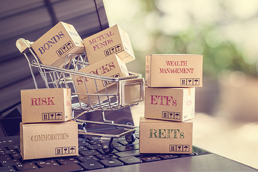 Small shopping cart placed on a computer keyboard holding boxes labelled bonds, stocks and mutual funds, with additional stacked boxes labelled ETFs, REITs, risk, commodities and wealth management in the foreground