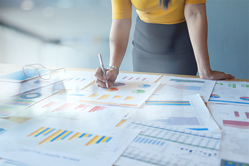 Person standing at a desk and writing on printed documents featuring charts and graphs, with additional data sheets spread across the surface and a pair of glasses placed nearby