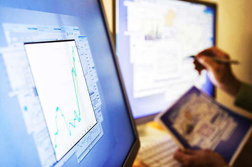 Close-up of two computer screens displaying charting software with line graphs and data points, with a person holding a tablet and using a stylus to interact with the display in a workstation setting