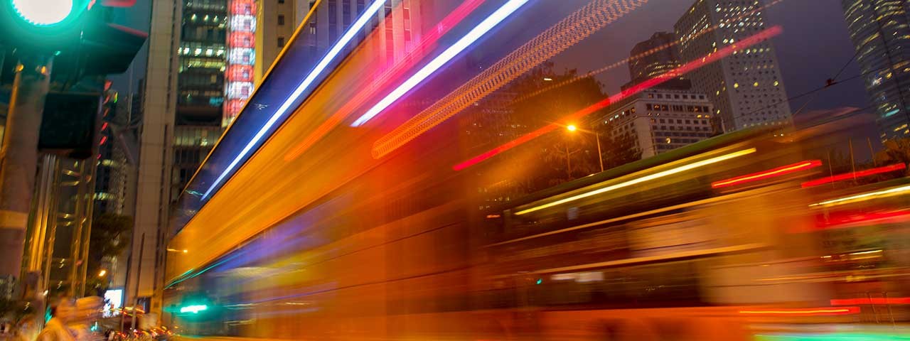 Long-exposure photograph of colourful light trails moving along a city street, with buildings and traffic lights visible in the background.