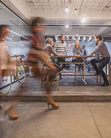 Group of people sitting and standing around a high table in a glass-walled office, while blurred figures walk past in the foreground and a bicycle is visible to the left.