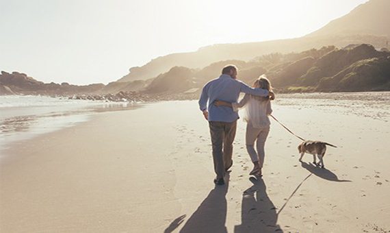 Outdoor scene of two people running along a beach with a dog near the shoreline and mountains in the background under soft natural light