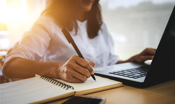 Close-up of a person writing in a notebook with a pen while using a laptop on a desk, with soft natural light illuminating the workspace