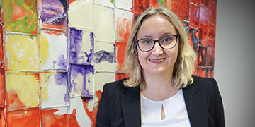 Head-and-shoulders professional portrait of a person wearing glasses and a blazer, positioned in front of a colourful abstract artwork background