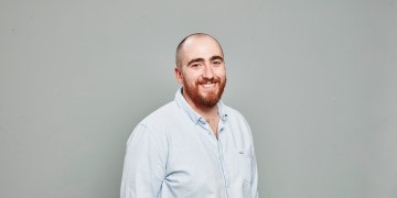 Head-and-shoulders professional portrait of a person with facial hair wearing a collared shirt, photographed against a plain light background