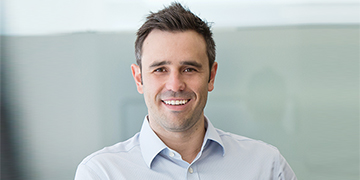 Head-and-shoulders professional portrait of a person wearing a collared shirt, facing the camera against a softly blurred indoor background