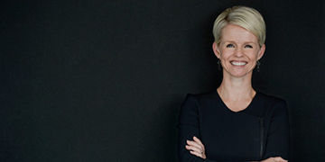 Head-and-shoulders professional portrait of a person wearing a dark top, positioned against a dark background with arms crossed