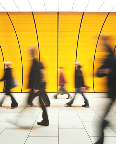 Blurred figures walking across a tiled floor in front of a large curved yellow wall with vertical lines.