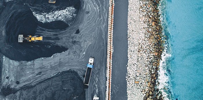Aerial view of a coastal industrial site with a dark excavation or stockpile area beside a narrow roadway, with the ocean on one side showing blue water and waves along a rocky shoreline