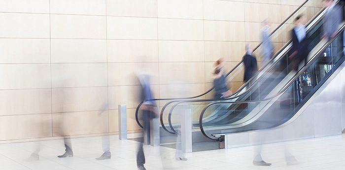 Blurred figures moving through a building lobby with an escalator and large wall panels.