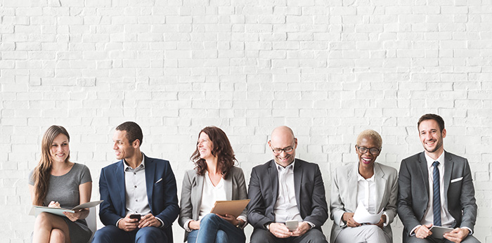 Six people seated in a row against a white brick wall, holding documents, tablets, and mobile devices.