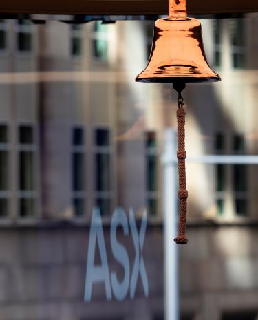A polished metallic bell with a rope pull hangs prominently in the foreground. Behind it, the letters "ASX" are clearly visible on a glass surface.