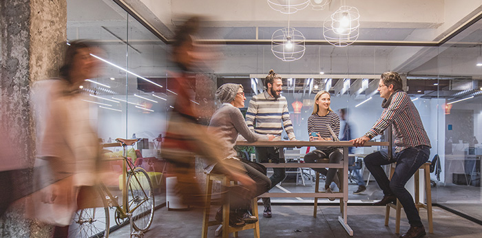 Group of people seated and standing around a table in a glass-walled office, with blurred figures walking past in the foreground and a bicycle visible nearby.