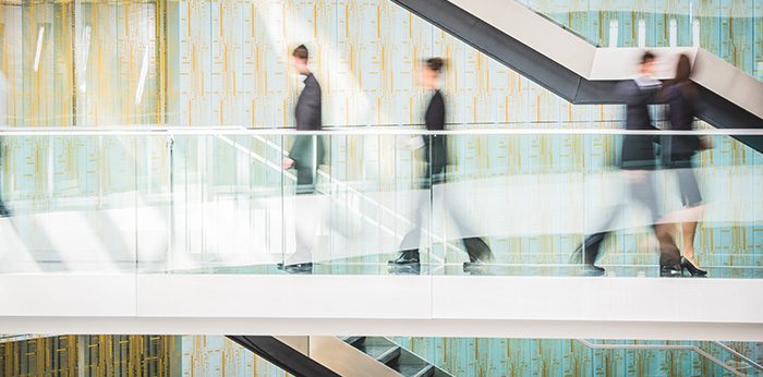 Blurred figures walking along elevated walkways and escalators inside a modern office building.