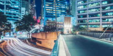 Blurred night-time city scene with illuminated office buildings, light trails from traffic, and a pedestrian walkway.
