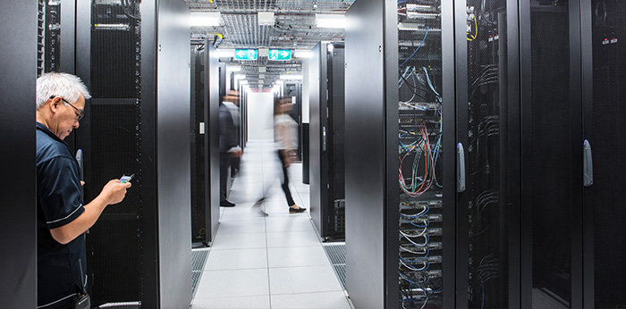 Data centre aisle with server racks on both sides, while people walk through the corridor and one person uses a mobile device.