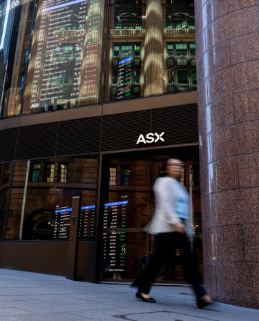 A person walks past the entrance of the ASX stock exchange building in an urban financial district. Large digital screens display market data and colourful charts reflected on the glass facade.