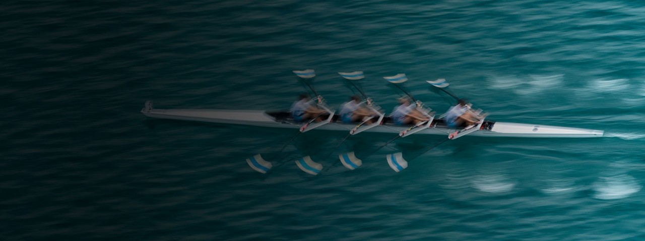 Aerial view of a long rowing boat with multiple rowers moving across green-blue water, leaving small ripples behind.